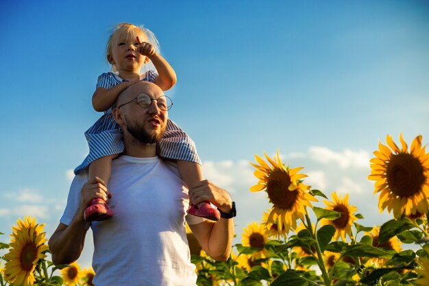 眼鏡をかけている若い男は、ひまわり畑を横切って彼の肩に子供を運びます。