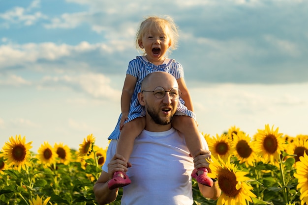 Young man wearing glasses holds a child on his shoulders on a sunflower field.