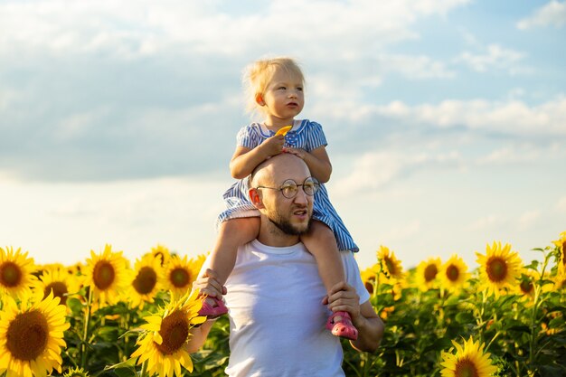 Young man wearing glasses holds a child on his shoulders on a sunflower field.