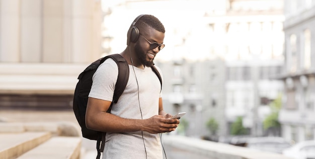 a young man wearing headphones and sunglasses uses his smartphone while walking through a city