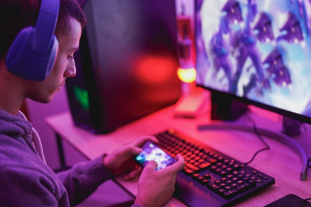 Photo young man wearing headset and play video games with smartphone online in front of computer