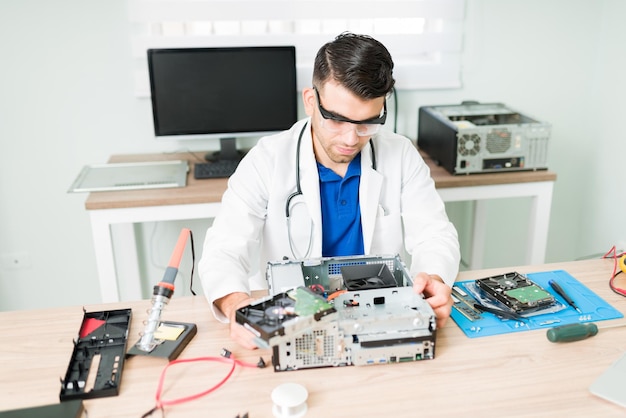 Giovane che indossa un camice da laboratorio e occhiali di protezione che riparano una cpu rotta. tecnico che lavora come medico informatico presso l'officina