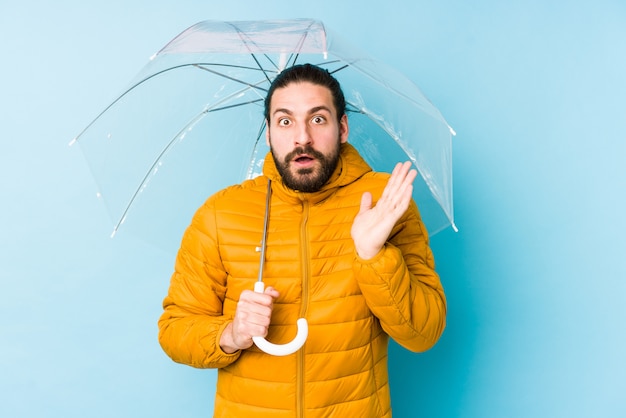 Young man wearing a long hair look holding an umbrella surprised and shocked.