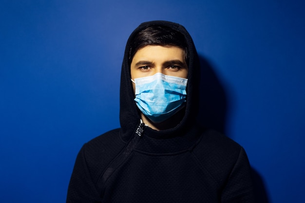 Молодой человек носить медицинскую маску гриппа и свитер с капюшоном. стена фантомно-синего цвета.