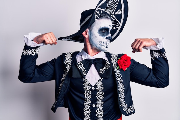 Young man wearing mexican day of the dead costume over white showing arms muscles smiling proud fitness concept
