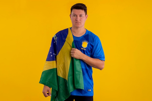 Young man wearing the official uniform shirt of the Brazilian soccer team in the 2022 Qatar Cup and with the flag of Brazil in studio photo Brazilian fan