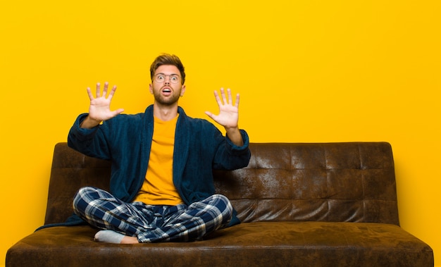 Young man wearing pajamas feeling stupefied and scared, fearing something frightening, with hands open up front saying stay away 