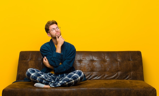 Young man wearing pajamas feeling thoughtful, wondering or imagining ideas, daydreaming and looking up to copy space . sitting on a sofa