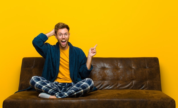Молодой человек в пижаме смеется, смотрит счастливым, позитивным и удивленным, понимая отличную идею, указывающую на боковое copyspace. сидя на диване
