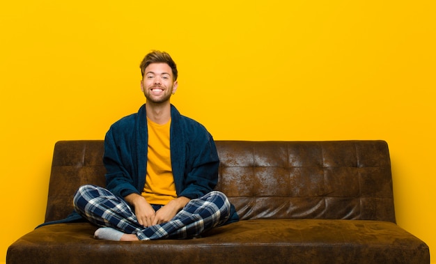 Young man wearing pajamas looking happy and goofy with a broad, fun, loony smile and eyes wide open . sitting on a sofa