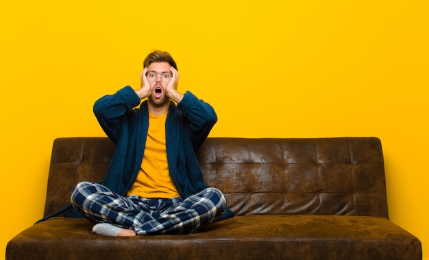 Young man wearing pajamas looking unpleasantly shocked, scared or worried, mouth wide open and covering both ears with hands . sitting on a sofa