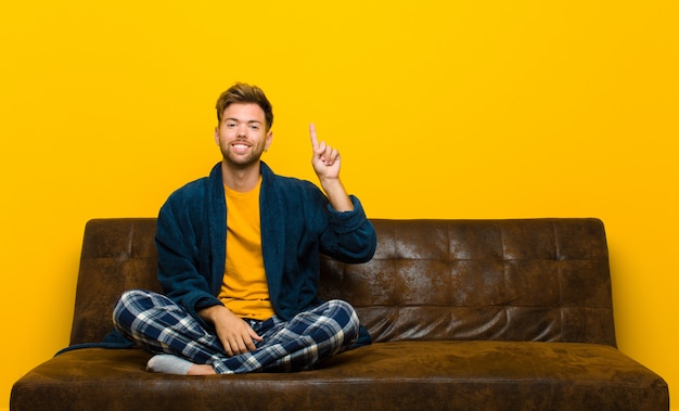 Young man wearing pajamas smiling cheerfully and happily, pointing upwards with one hand to copy space . sitting on a sofa