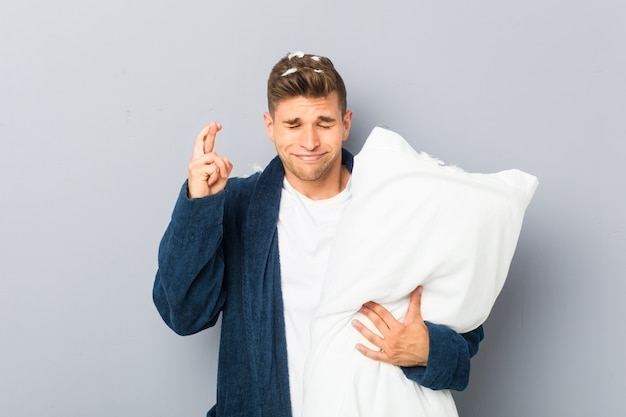 Young man wearing pijama holding a pillow crossing fingers for having luck