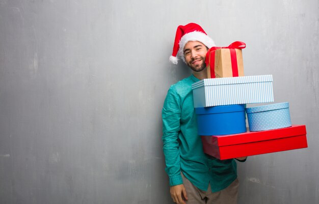 Young man wearing a santa hat holding gifts crossing arms, smiling and relaxed