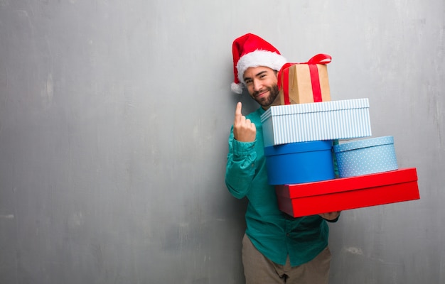 Young man wearing a santa hat holding gifts inviting to come