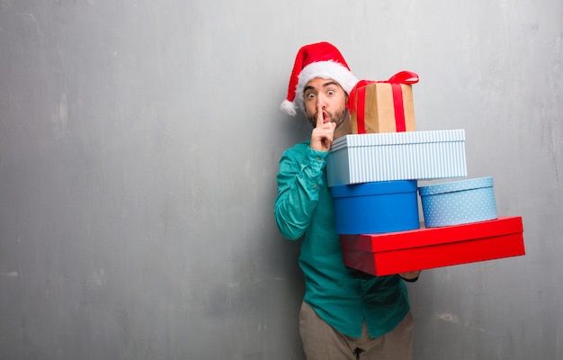 Young man wearing a santa hat holding gifts keeping a secret or asking for silence