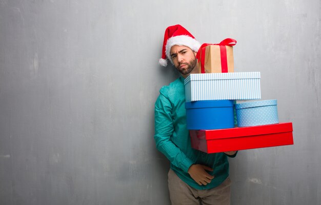 Молодой человек в шляпе Санта держит подарки, глядя прямо вперед