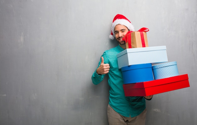 Молодой человек в шляпе Санта держит подарки, улыбаясь и поднимая пальца вверх