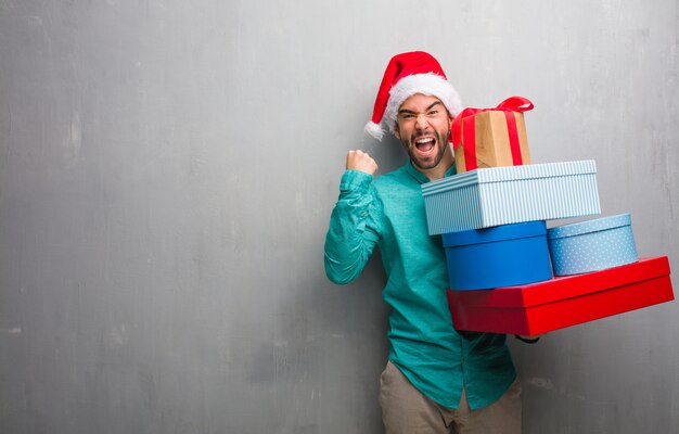 Young man wearing a santa hat holding gifts surprised and shocked