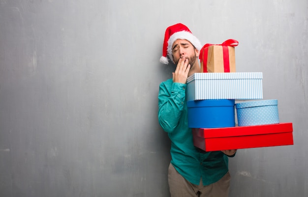 Photo young man wearing a santa hat holding gifts tired and very sleepy