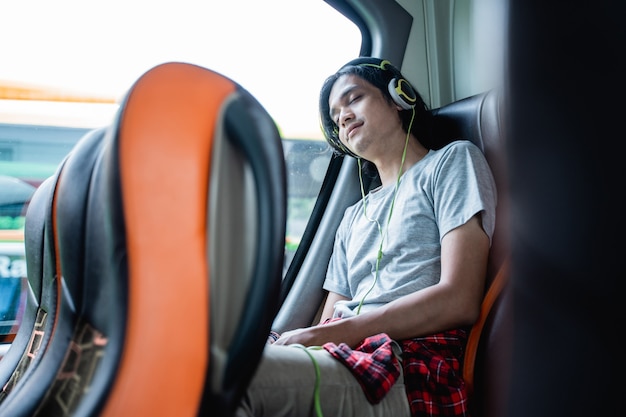 A young man wearing sleep headphones is leaning against the window while sitting by the window on a bus trip