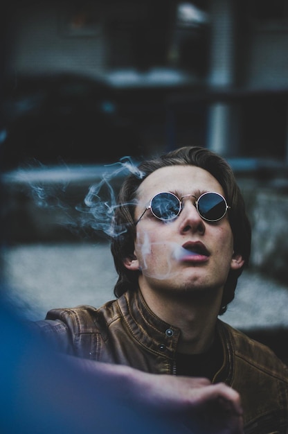 Photo young man wearing sunglasses and leather jacket while emitting smoke from mouth