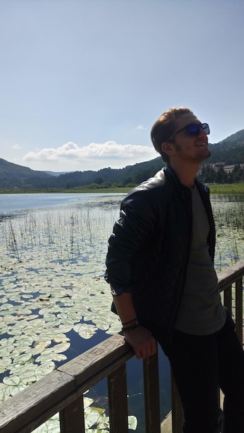Foto giovane con gli occhiali da sole in piedi accanto alla ringhiera contro il lago
