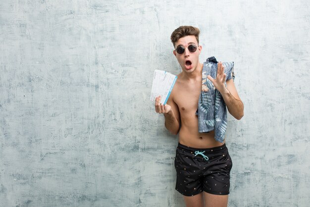 Young man wearing a swimsuit holding air tickets shouts loud, keeps eyes opened and hands tense.