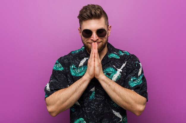 Young man wearing a vacation look holding hands in pray near mouth, feels confident.