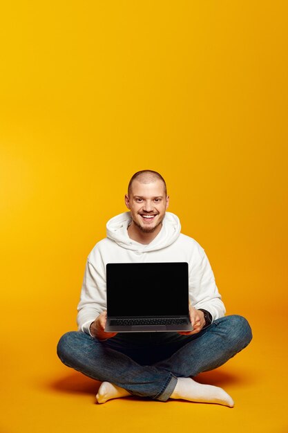 Young man wearing white hoodie smiling and showing new laptop in hands while sitting on floor