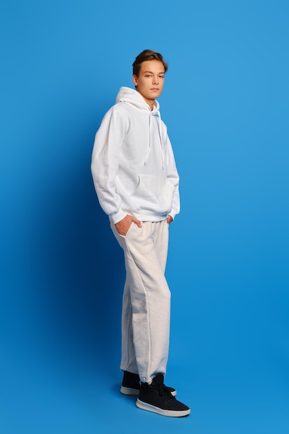 Young man wearing white sweatshirt and sweatpants standing over blue studio background with hands in pockets