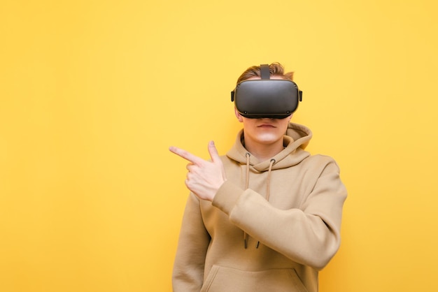 A young man wears a VR helmet and points his finger away at a blank space isolated on a yellow background wears casual clothes Teen guy uses VR glasses and points his finger at Copy space