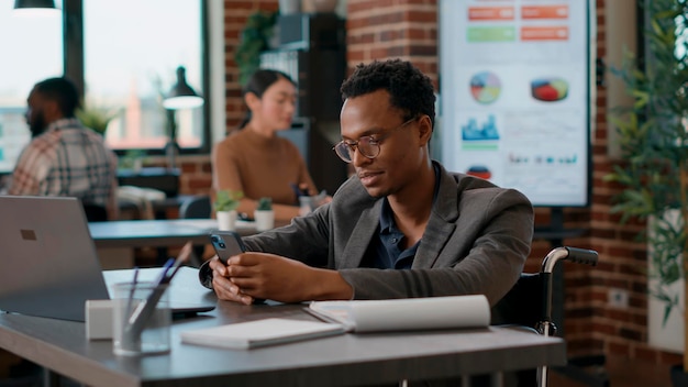 Young man in wheelchair browsing internet on smartphone, searching financial strategy for business sales growth. Person working with mobile phone in disability friendly office.