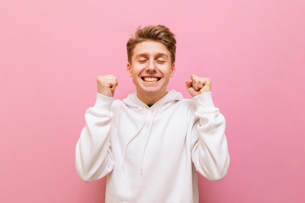 young man in white casual clothes rejoices with closed eyes and raised hands on pink background