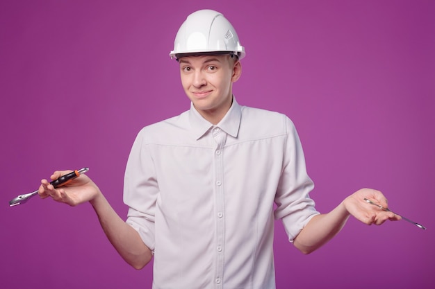 Young man in white helmet with working tool in hand on purple background don't know what to do