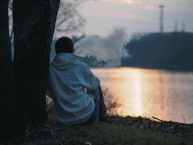 A young man in a white hoodie sits on the riverbank at sunset and exhales smoke from an electronic cigarette Vaping man