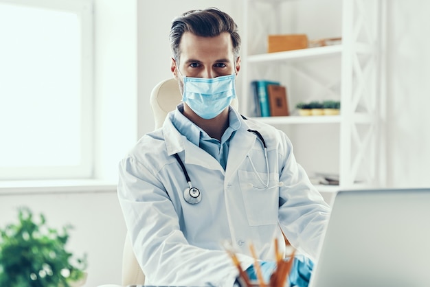 Young man in white lab coat and protective mask looking at camera while sitting indoors