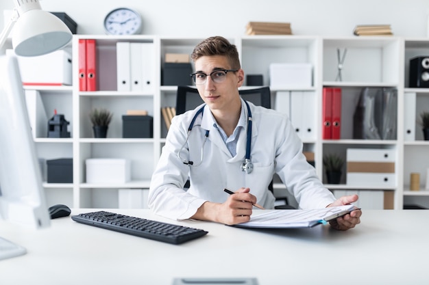 A young man in a white robe sitting at a table in the office. He holds a pen in his hand and looks straight. The portrait of the doctor.