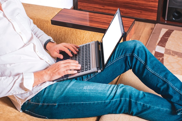 Young man in white shirt and blue jeans working on laptop, writes in notebook, looking on white screen in home office sitting on cozy beige sofa