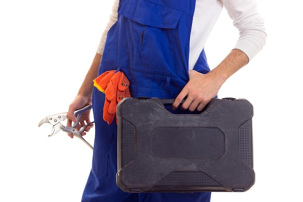 Young man in white shirt and blue overall with orange gloves holding spanner and black toolbox on white background in studio