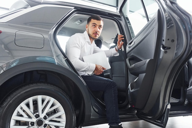 Young man in white shirt is indoors with modern new automobile