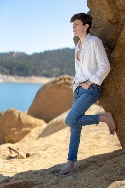 Молодой человек в белой рубашке и джинсах, прислонившись к скале