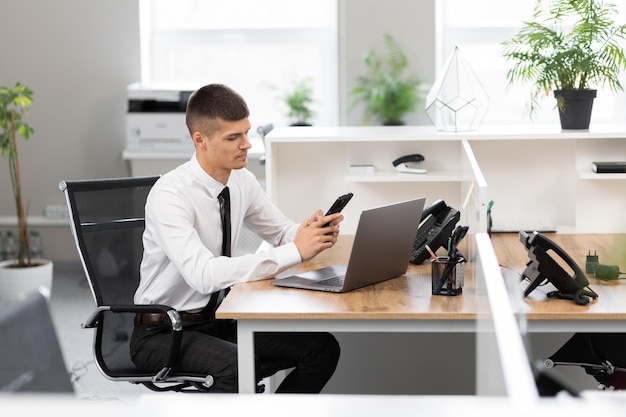 Young man in a white shirt with a black tie at the desk at the bright office working on a laptop