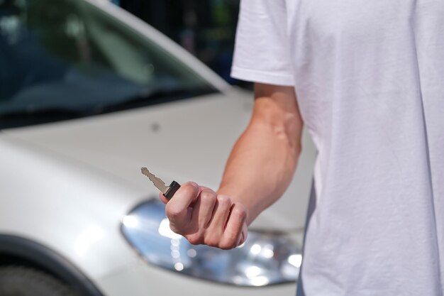 young man in a white t-shirt holds and shows a car key against the background of a white car