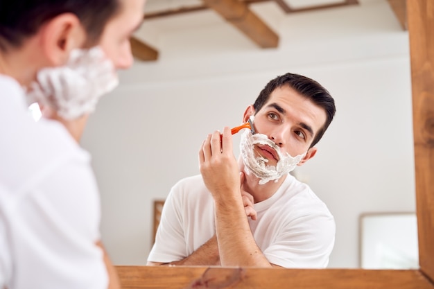 Young man in white T-shirt shaves while standing near mirror in bath in morning