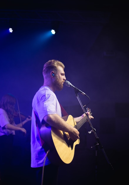 A young man in a white T-shirt with a beard holds an acoustic guitar in his hand and sings into a microphone stands on the stage