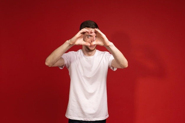 Молодой человек в белой футболке на красном фоне с синдромом Тричера показывает руками свое сердце людям с генетическими заболеваниями с ограниченными возможностями