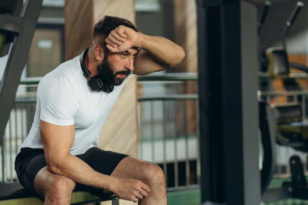 A young man wipes sweat from his forehead in the gym He is tired and exhausted from weight lifting and strength training