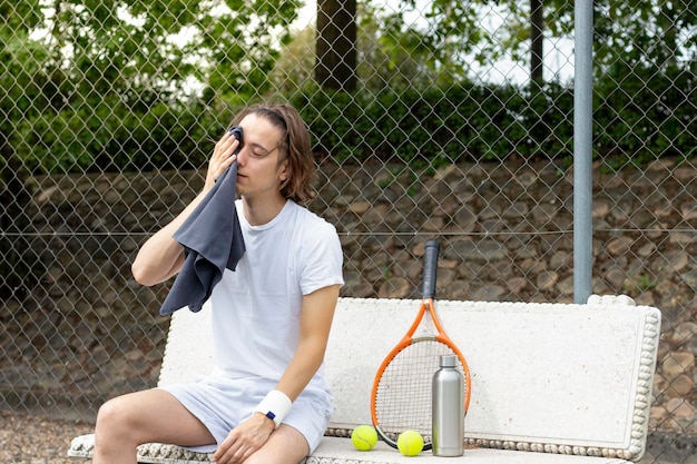 Young man wiping his sweat with a towel sitting on a bench after playing a game of tennis on a court