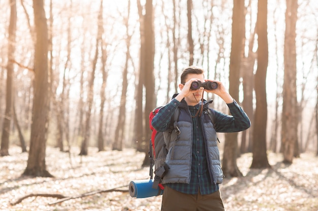 Young man with backpack looking at the binoculars, hiking in the forest